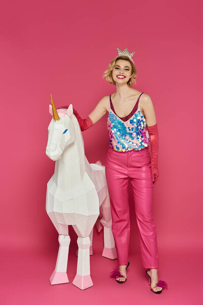Woman in pink sparkly outfit poses with white unicorn on pink background.