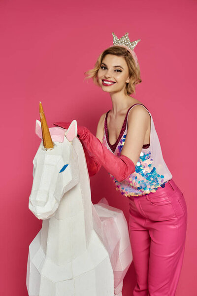 Woman in pink clothing and crown smiles by white unicorn on pink background.