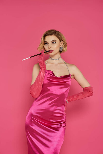 Женщина в розовом атласном платье и перчатках держит держатель для сигарет, позируя на розовом фоне.