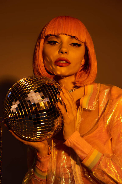 A woman with pink hair and a vibrant outfit holds a disco ball.