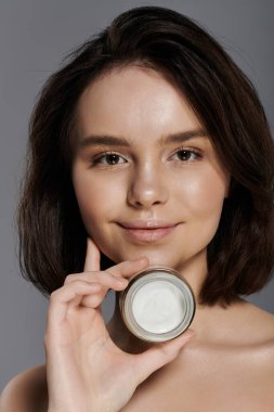 A woman with flawless skin holds a jar of cream in front of her face, smiling gently. clipart