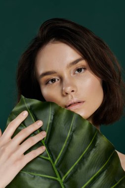 Woman with short brown hair poses against green backdrop, holding large green leaf. clipart