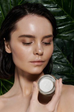 Bir kadın bir kavanoz kremayı canlı yeşil yaprakların arka planına karşı tutuyor..