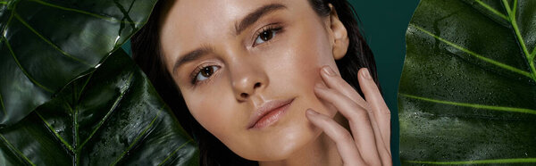 Портрет крупным планом женщины с рукой на щеке, обрамленной яркими зелеными листьями.