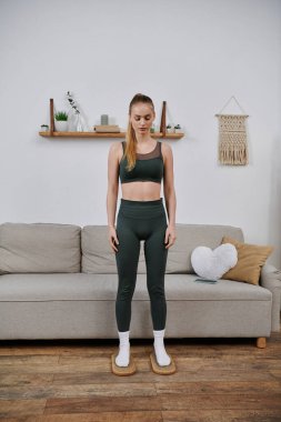 Yeşil spor elbiseli genç bir kadın oturma odasında duruyor, gözleri düşüncelere dalmış gibi kapalı..