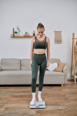 Spor kıyafetleri giyen bir kadın evinde bir terazide duruyor..