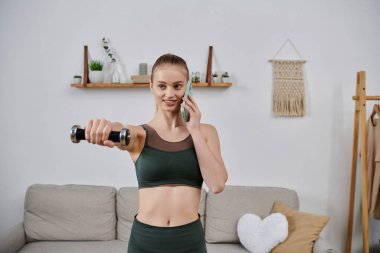 Atletizmde genç bir kadın evde telefonla konuşurken halterle egzersiz yapıyor..