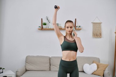 Spor kıyafetli bir kadın elinde bir dambıl tutup telefonuyla konuşuyor..