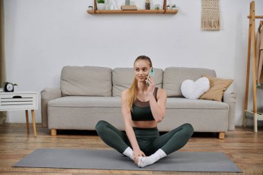 Oturma odasında yoga minderinde egzersiz kıyafetleri giymiş bir kadın, antreman sırasında telefon etmek için duraklıyor..