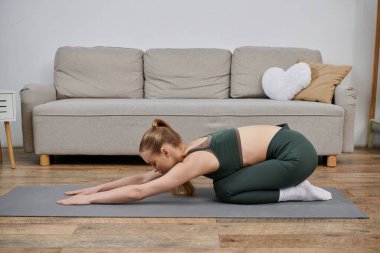 Genç bir kadın evinde yoga yapıyor. Kanepenin önünde gri bir paspasın üzerinde diz çöküyor..