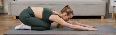 Yeşil spor kıyafetli genç bir kadın evdeki mindere yoga pozu veriyor..