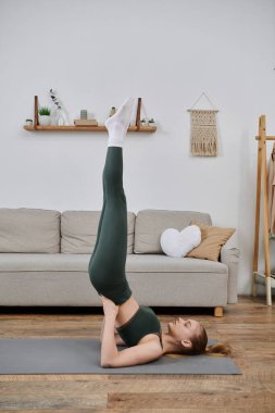 Genç bir kadın evde yoga yapıyor, paspasın üstünde ters pozlar veriyor..