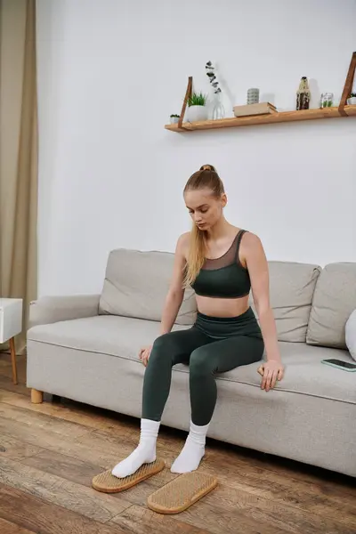 stock image A young woman in athletic wear sits on a couch at home, using foot massage tools to relax.