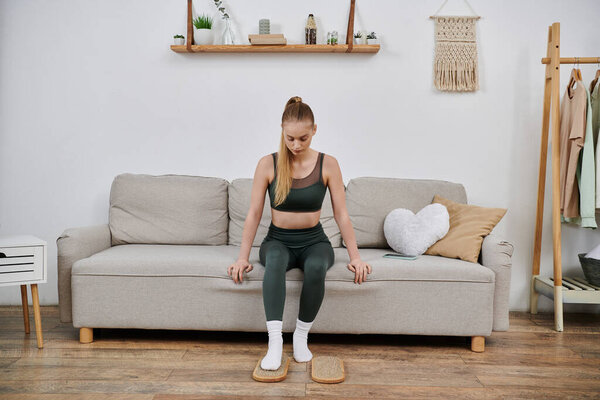 A young woman in athletic wear sits on a couch, using a foot massage tool at home.