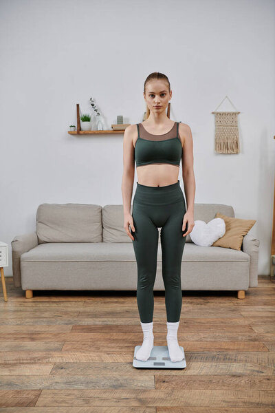 A young woman in athletic wear stands on a scale in her living room.