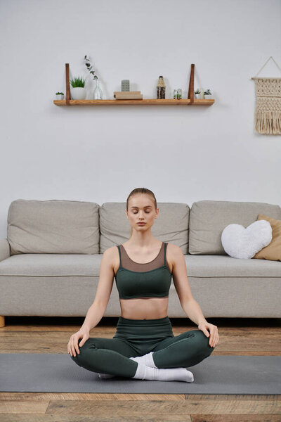 Молодая женщина практикует йогу дома, сидя в медитативной позе на сером коврике.