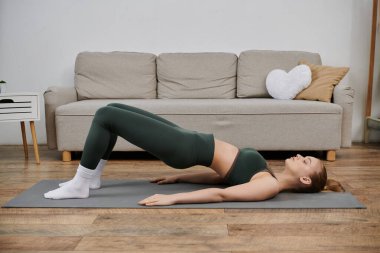 Yeşil spor elbiseli genç bir kadın oturma odasındaki yoga minderinde köprü pozu veriyor..