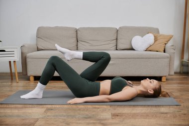 Evde yoga minderinde egzersiz yapan genç bir kadın sırt üstü yatarken bacağını kaldırıyor..