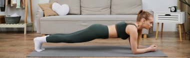 Genç bir kadın oturma odasındaki yoga minderinde tahta egzersizi yapıyor..