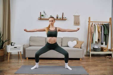 Yeşil spor sutyen ve taytlı genç bir kadın oturma odasında yoga pozu veriyor..