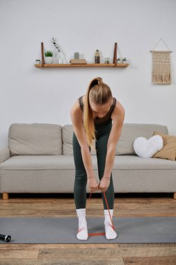 Antrenman elbiseli bir kadın oturma odasındaki yoga minderinde kırmızı bir direniş bandıyla esniyor..