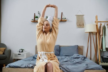 A young woman in a robe stretches on a bed in her home. clipart