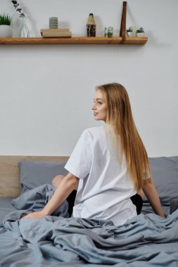 Genç bir kadın yatağında oturuyor, kameradan kafasını çeviriyor, düşüncelere dalmış..
