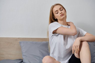 Genç bir kadın evinde bir yatakta oturuyor, omuzlarını geriyor..