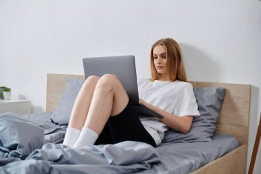 Genç bir kadın rahat bir şekilde yatağında oturuyor, dizüstü bilgisayarına dalmış..