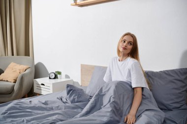 Genç bir kadın yatağında oturmuş, düşünceli bir şekilde çerçevenin dışındaki bir şeye bakıyor..