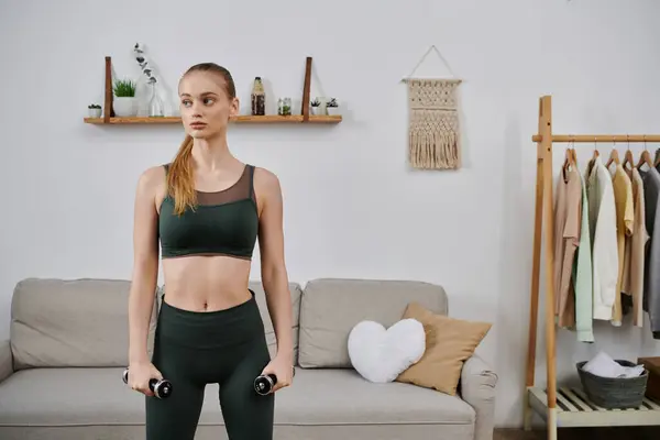 stock image A young woman in athletic wear stands in her living room holding dumbbells.