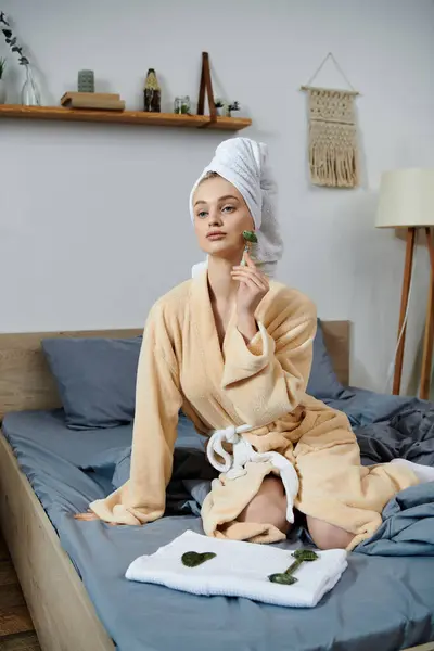 stock image A young woman in a bathrobe uses a jade roller on her face while sitting on a bed.