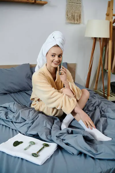 Stock image A young woman sits on a bed in a robe, holding a jade roller and smiling.