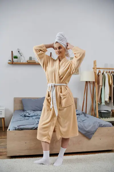 stock image A young woman in a robe and towel on her head stands by her bed, ready to start her day.