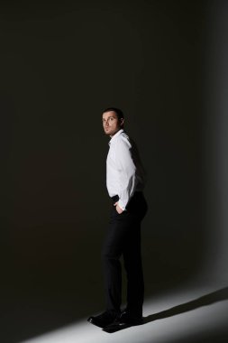 A man in a white shirt and black pants stands in a spotlight, looking away from the camera. clipart