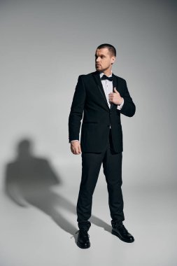 Confident man in tuxedo adjusts collar against gray backdrop. clipart