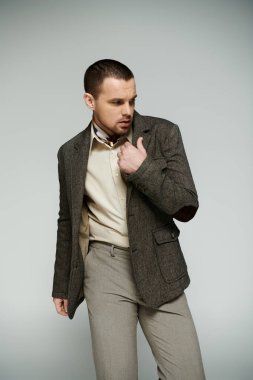 Tüvit ceketli şık bir adam gri bir fonda duruyor, yakasını düzeltiyor..