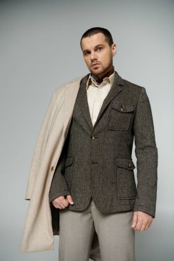 Tüvit ceketli ve bej pantolonlu bir adam gri bir fonda duruyor..