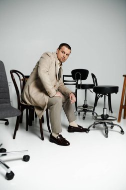 A stylish man sits on a chair, surrounded by other chairs in a studio setting. clipart