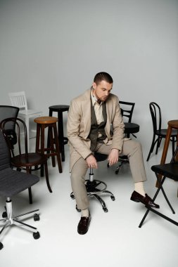 A man in a beige suit sits on a swivel chair surrounded by other chairs. clipart