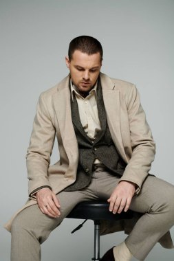 A stylish man in a beige coat, vest, and trousers sits on a stool, looking down thoughtfully. clipart