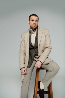 A stylish man wearing a beige coat and brown shoes sits on a stool against a gray backdrop. clipart