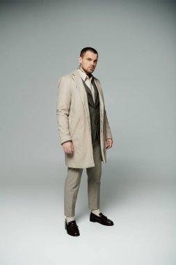 Krem rengi trençkotlu bir adam açık gri bir arka planda duruyor ve doğrudan kameraya bakıyor..
