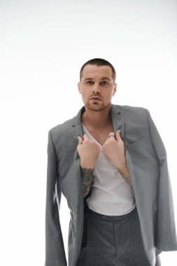 Gri takım elbiseli bir adam stüdyoda duruyor, kendine güvenen bir ifadeyle ceketini düzeltiyor..