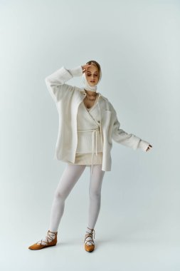 Beyaz elbiseli genç bir kadın stüdyoda duruyor, elini yüzüne kaldırıyor..