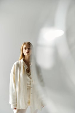 Beyaz elbiseli genç bir kadın parlak bir stüdyoda duruyor, yumuşak, ruhani bir ışıkla aydınlatılıyor..