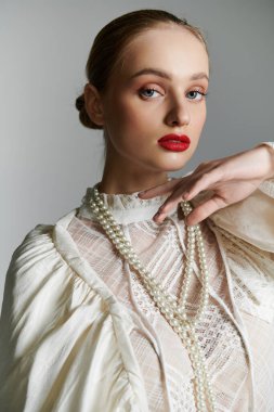 Beyaz bluzlu genç bir kadın, inci kolyeli, arka planda poz veriyor..