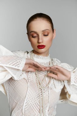 Beyaz bluzlu genç bir kadın inci kolye takar, derin derin düşünür..