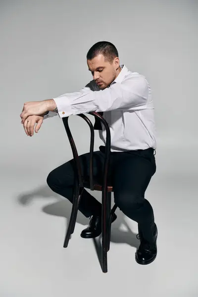 stock image A man in a white shirt and black trousers sits on a chair, leaning back with his arms crossed.