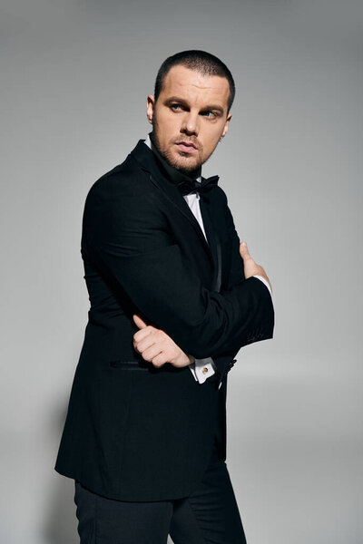 Человек в черном смокинге стоит на сером фоне, руки скрещены, смотрит в сторону.